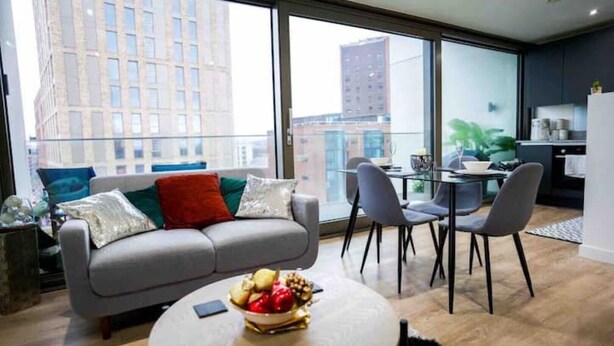 Ein modernes Wohnzimmer mit einem grauen Sofa, einem runden Couchtisch mit Obst und einem Essbereich mit Stühlen und einem Glastisch.