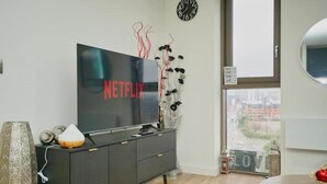 Ein modernes Wohnzimmer mit einem großen Flachbildfernseher, einem schlichten Schrank, einer Zierpflanze, einer Uhr und Blick auf die Stadt durch ein großes Fenster.
