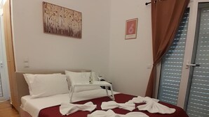 A neatly made bed with a red bedspread and white pillows, a small bedside table with a bottle and glass, and a window with blinds. - Grandma’s Pine Apartments – Cozy Double Bed (Paralia Politikon)