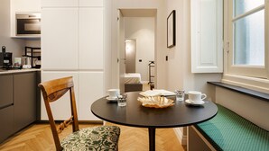 A compact kitchen with a built-in oven and coffee maker, a dining area with a round table and bench, and a view of a bedroom through an open door. - Corvetto Luxury Suite by Wonderful Italy (Genova)