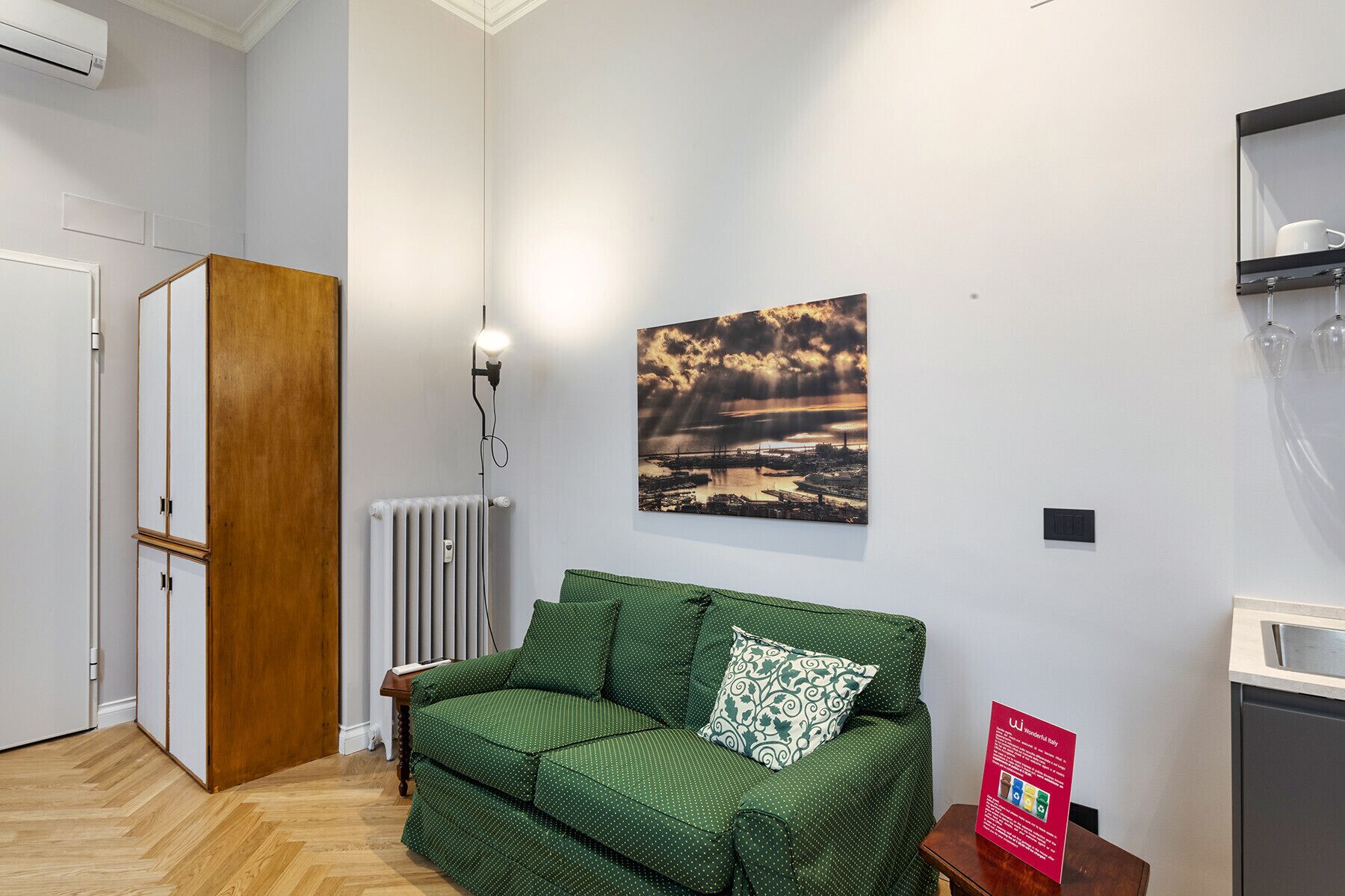 A room with a green sofa, a radiator, a wall-mounted picture, and a small table with a sign.