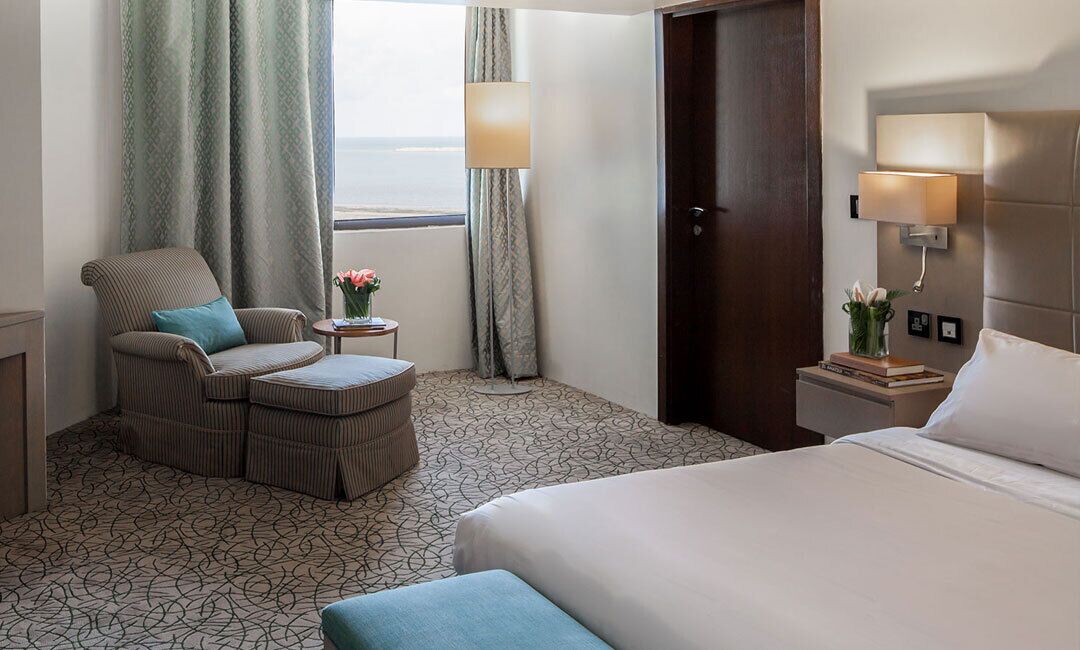 A hotel room with a bed, a chair with a cushion, a small table with a vase of flowers, a lamp, and a view of the sea through a window.