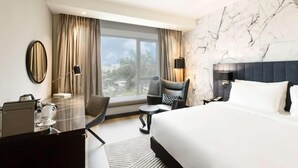 A hotel room with a large bed, a desk with a coffee machine, a chair, a small table, and a view of palm trees through the window.