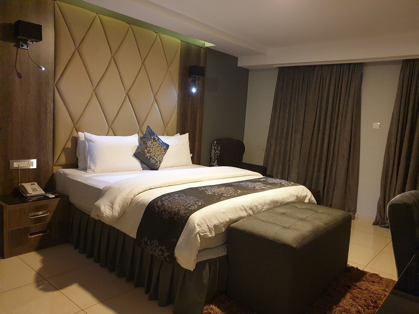 A hotel room with a large bed, a bedside table with a phone, a chair, and a curtain.