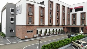 A hotel with a modern design, featuring a mix of brick and white exterior, multiple balconies with plants, and a parking area in front.