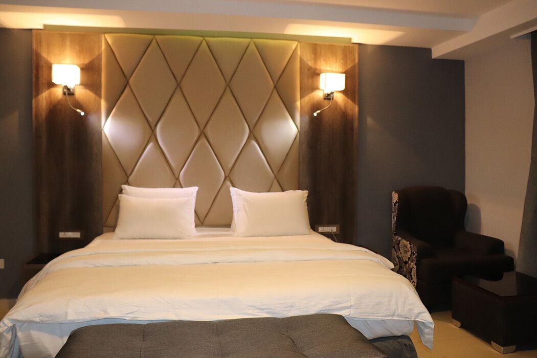 A hotel room with a large bed, two bedside lamps, a headboard with a diamond pattern, and a dark armchair.