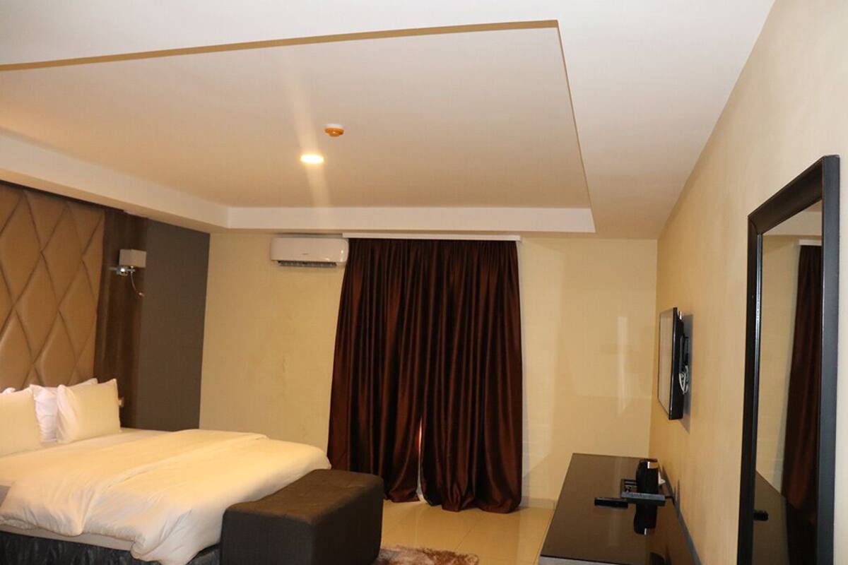 A hotel room with a bed, a desk, a chair, a mirror, a window with curtains, and an air conditioning unit.