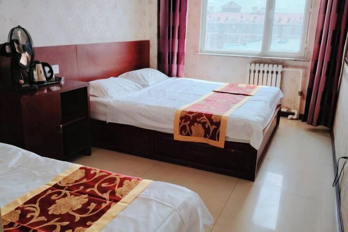 Habitación de hotel con dos camas, una mesita de noche de madera, una tetera y un ventanal con cortinas.