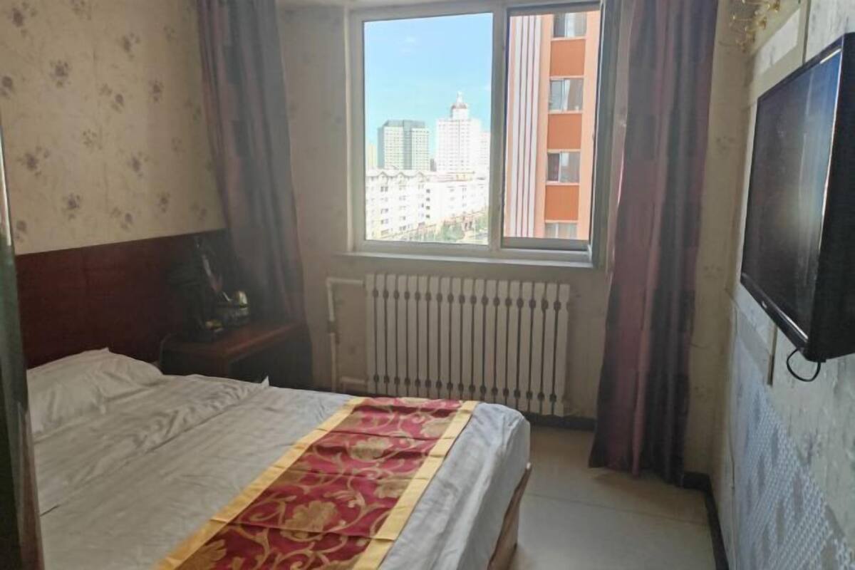 Una habitación de hotel con cama, ventana con vista a edificios, radiador y televisor en la pared.