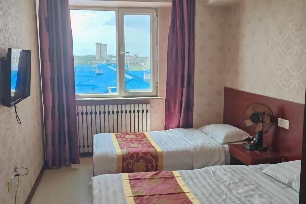 Habitación de hotel con dos camas, una ventana con cortinas, un radiador, un televisor en la pared y un ventilador en la mesita de noche.