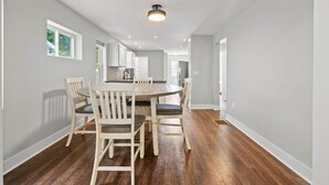 A spacious dining area with a round table and six chairs, a kitchen in the background, and a ceiling light fixture.