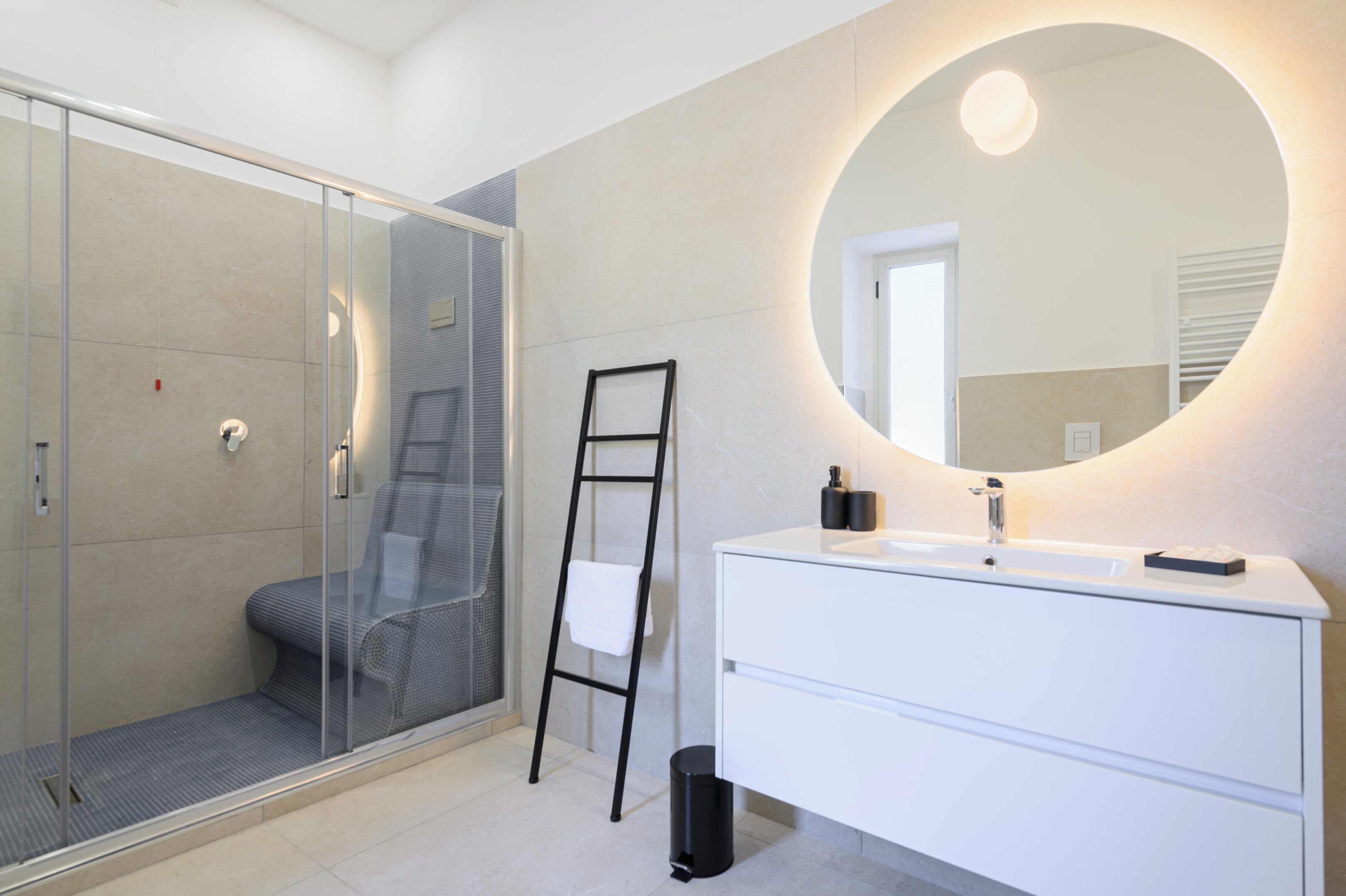 A modern bathroom with a glass shower enclosure, a white vanity with a round mirror, and a black trash bin.