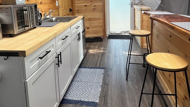 Una cocina compacta con muebles de madera, horno empotrado y un lavamanos. Hay dos taburetes y una pequeña barra.