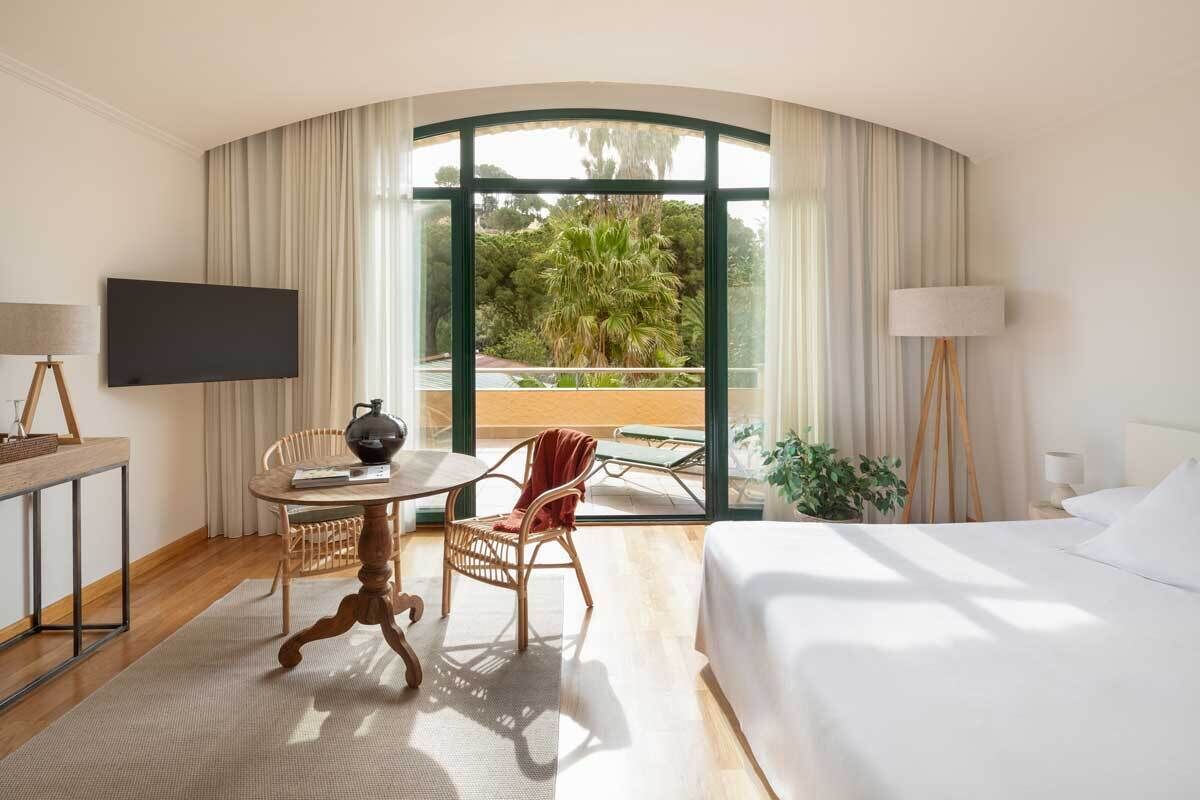 A hotel room with a curved glass wall, a bed, a desk with a flat-screen TV, a wicker chair, a small table with a teapot, and a potted plant.