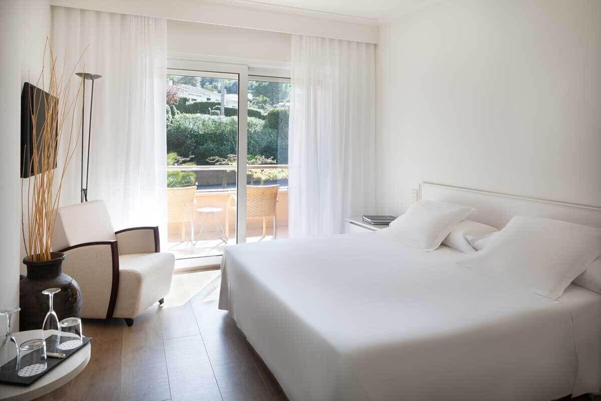 A hotel room with a large bed, a chair, a small table with glasses, and a view of a balcony with greenery.