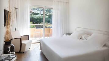 A hotel room with a large bed, a chair, a small table with glasses, and a view of a balcony with greenery.