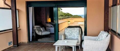 A balcony with a white table and two wicker chairs, a sliding glass door leading to a room with a bed and a lamp, and a view of the landscape.