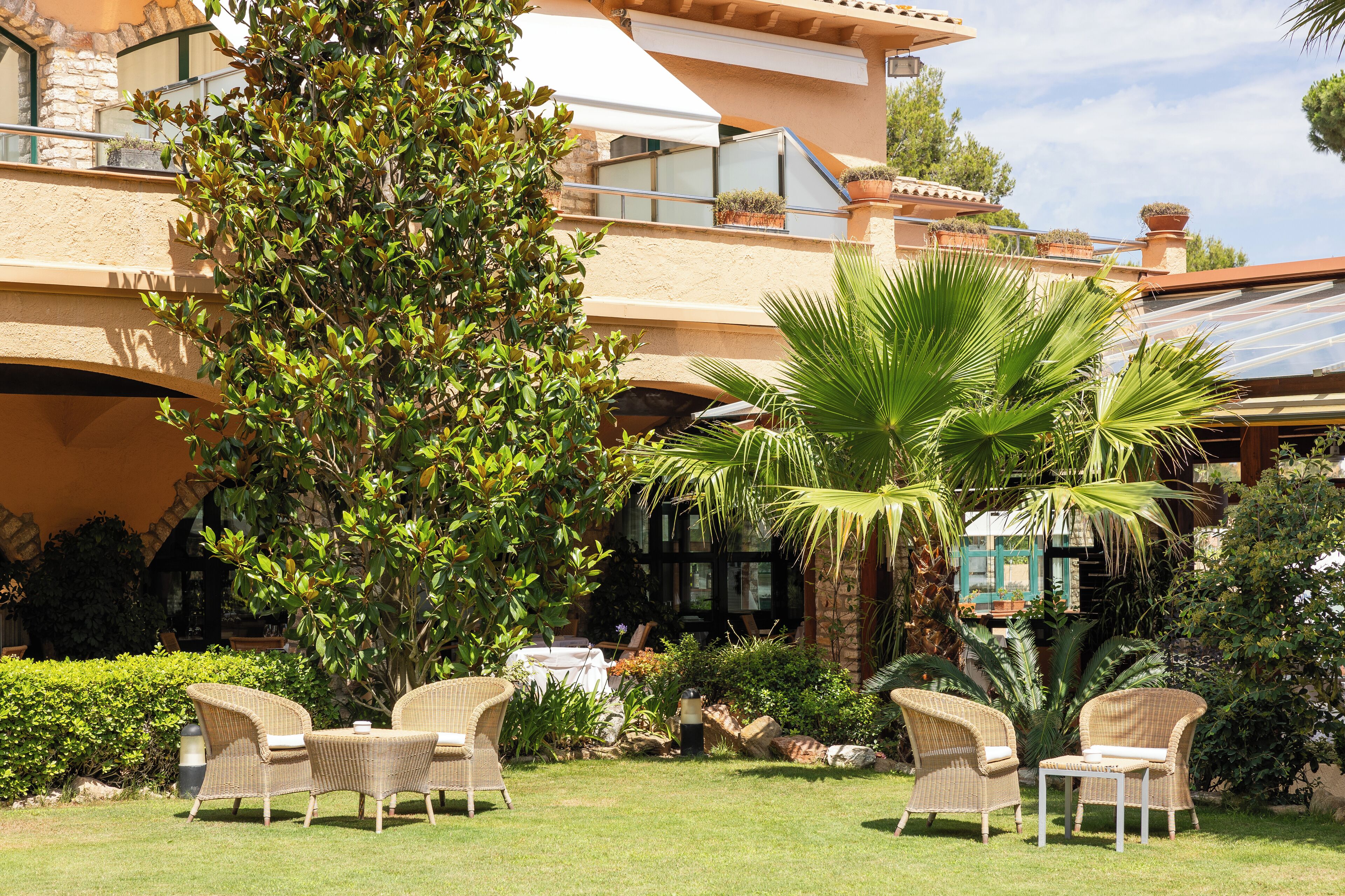 A well-maintained outdoor patio area with wicker furniture, lush greenery, and a stone building in the background.