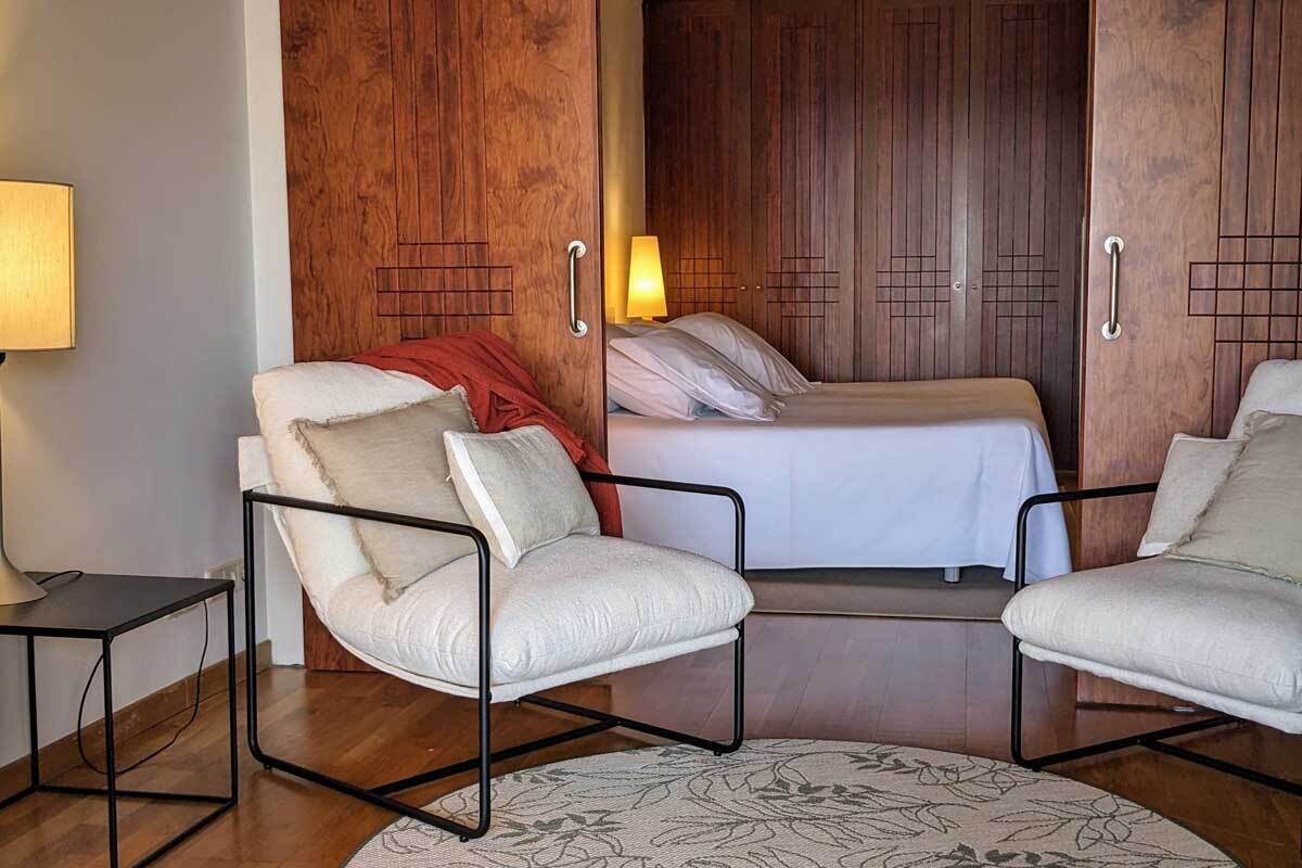 A hotel room with two single beds, a wooden wardrobe, a small table with a lamp, and two armchairs.