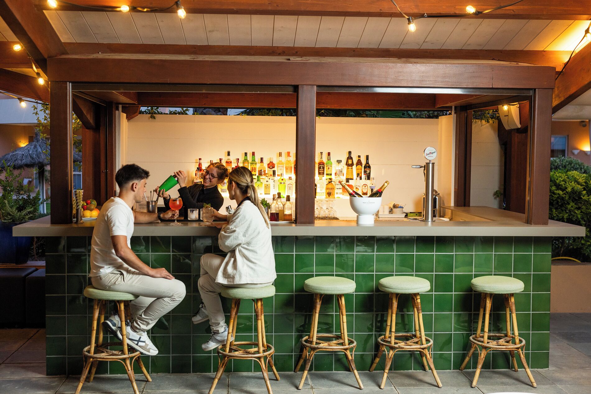 A bar area with a green tiled wall, wicker bar stools, and a variety of bottles and glasses on the counter.