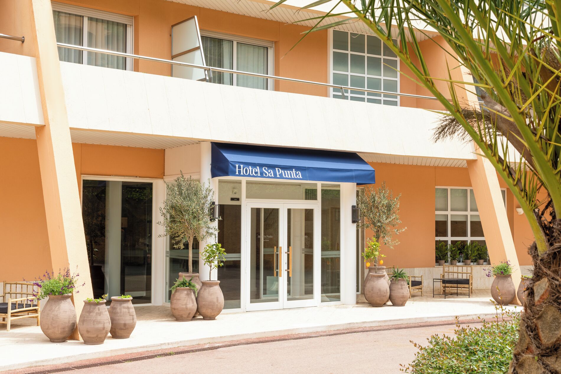 A hotel entrance with a blue awning, large potted plants, and a seating area with chairs and a table.