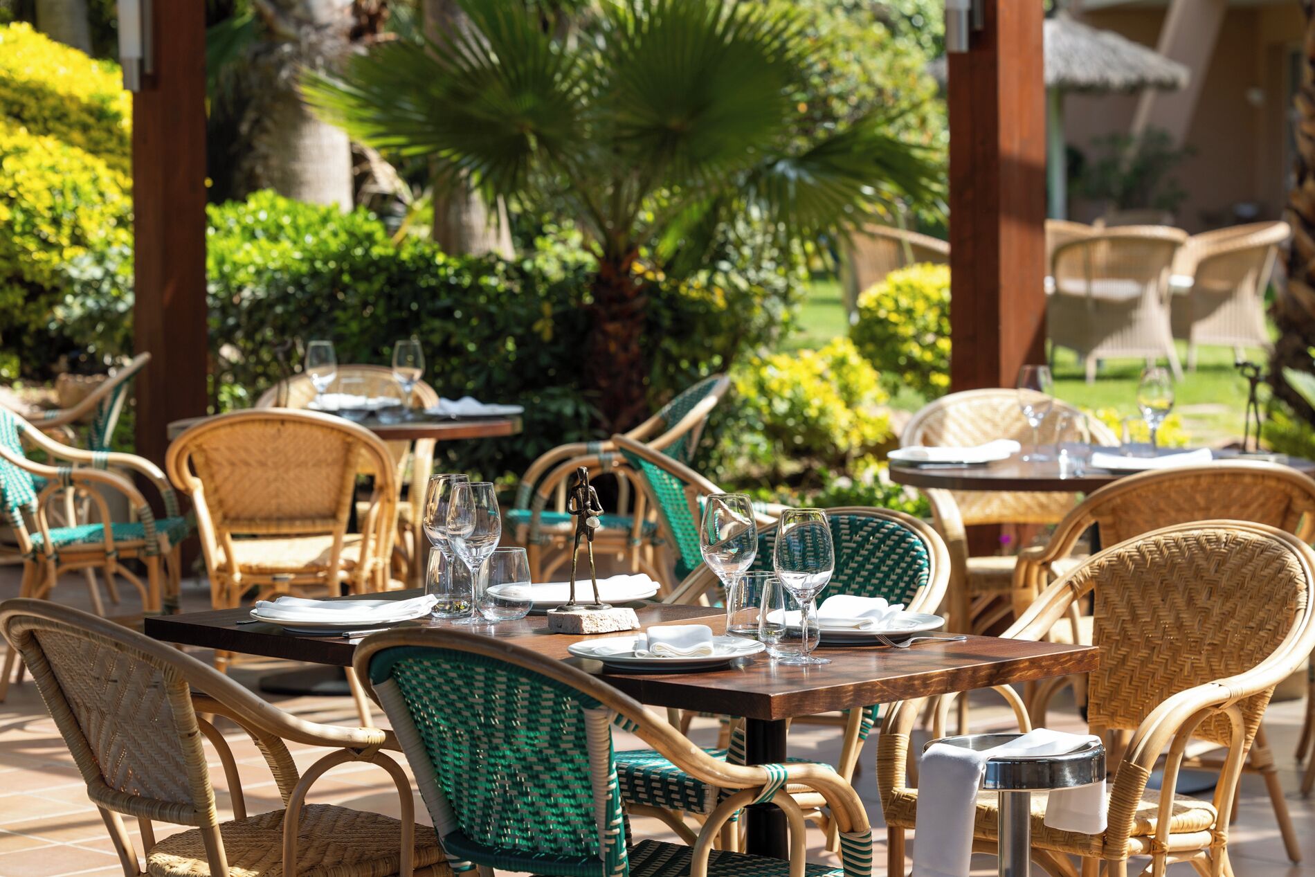 A restaurant patio with wicker chairs and tables, set in a lush garden with palm trees and greenery.