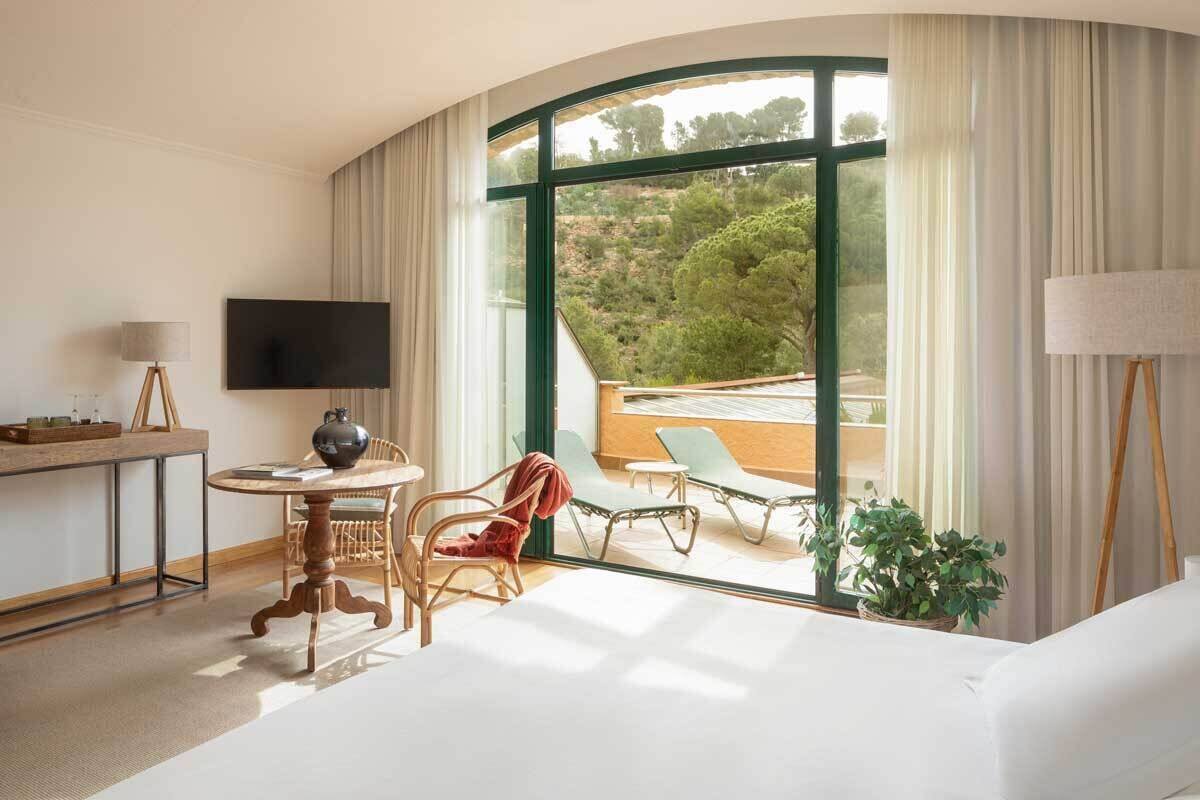 A hotel room with a large window, a bed, a desk with a TV, and a chair.