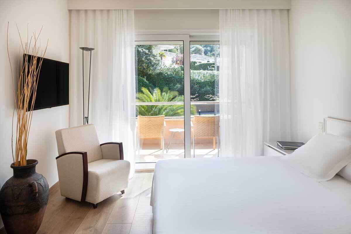 A hotel room with a bed, a chair, a TV, a vase with branches, and a balcony with a table and chairs.