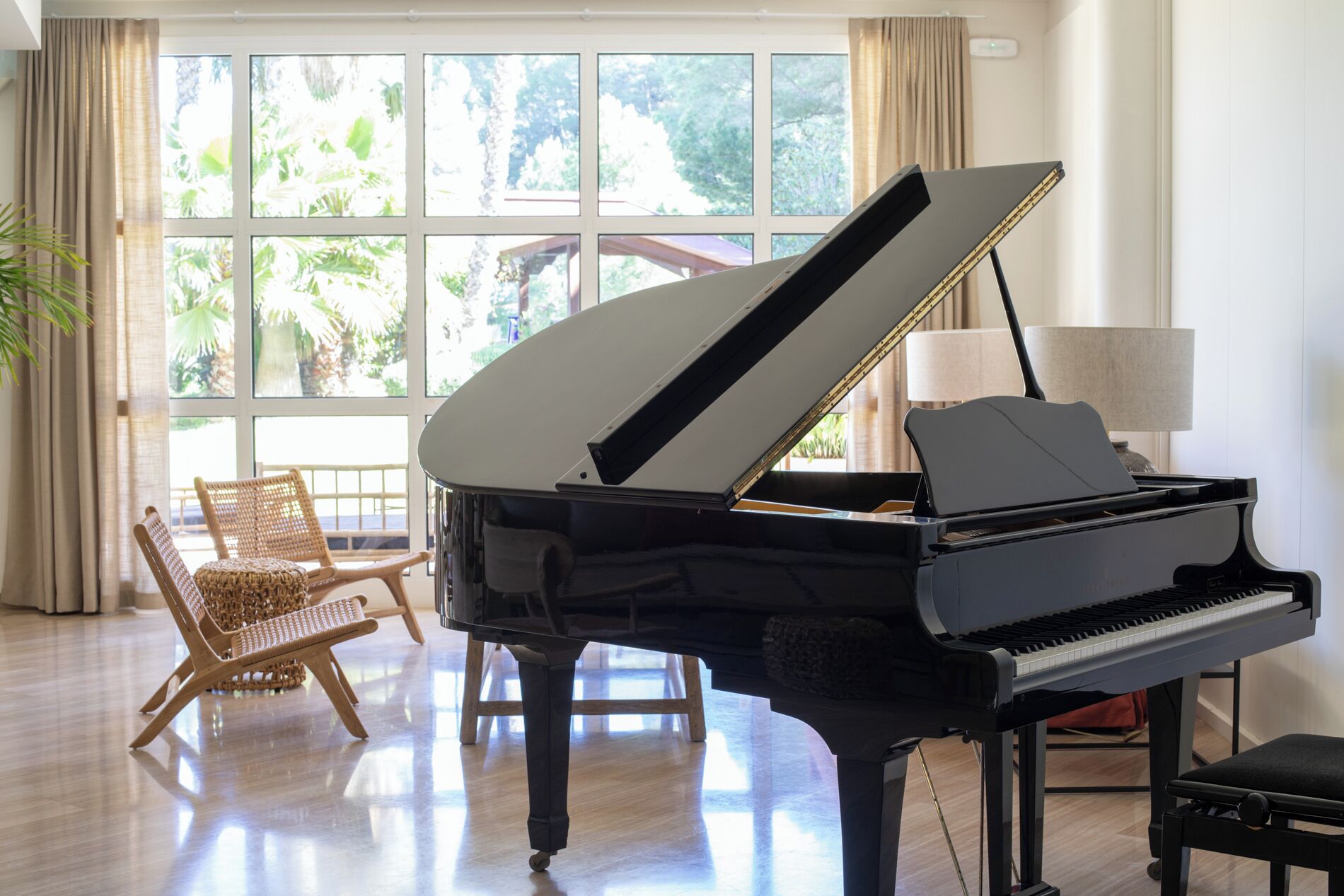 Un piano à queue dans une pièce bien éclairée, dotée de grandes fenêtres, d’un fauteuil en osier et d’une lampe.