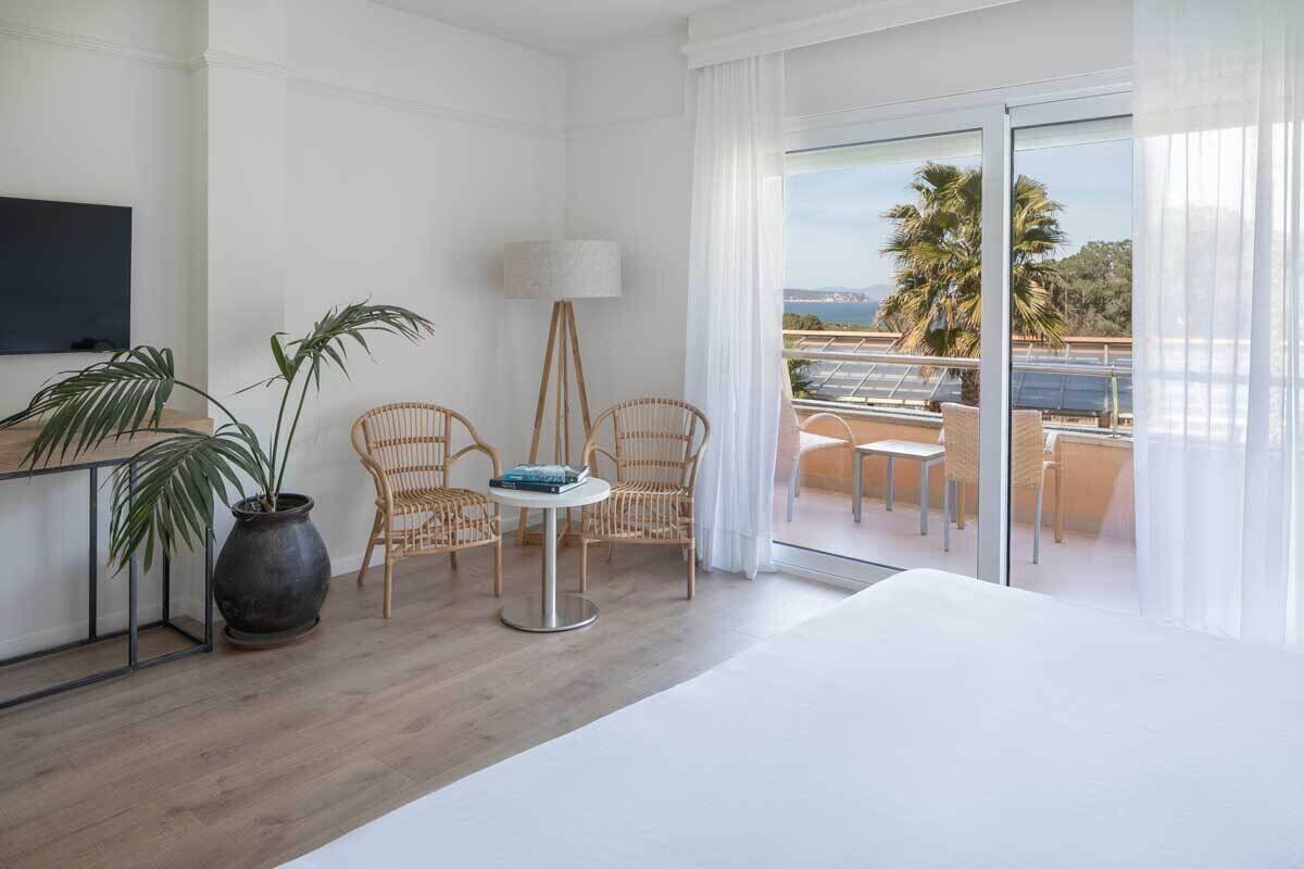 A hotel room with a balcony, a bed, a table with chairs, and a potted plant.