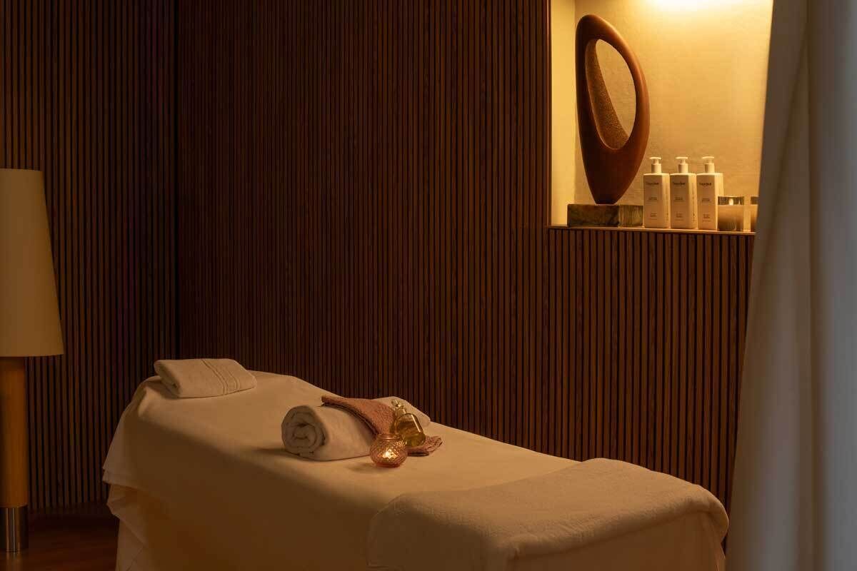 Een massagetafel met handdoeken en decoratie, een lamp en een plank met flessen in een kamer met houten lambrisering.