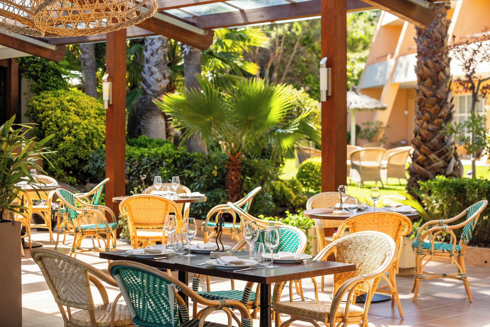 Un espacio exterior para comer con muebles de mimbre y mesas dispuestas para dos, rodeado de abundante vegetación y palmeras.