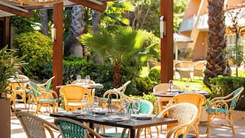 Un espacio exterior para comer con muebles de mimbre y mesas dispuestas para dos, rodeado de abundante vegetación y palmeras.