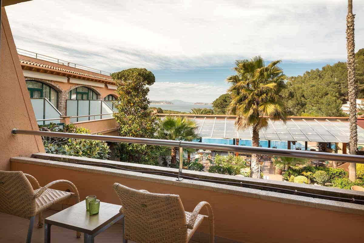 Un balcón con sillas de mimbre y una mesa, con vista a una zona de piscina con palmeras y al mar.
