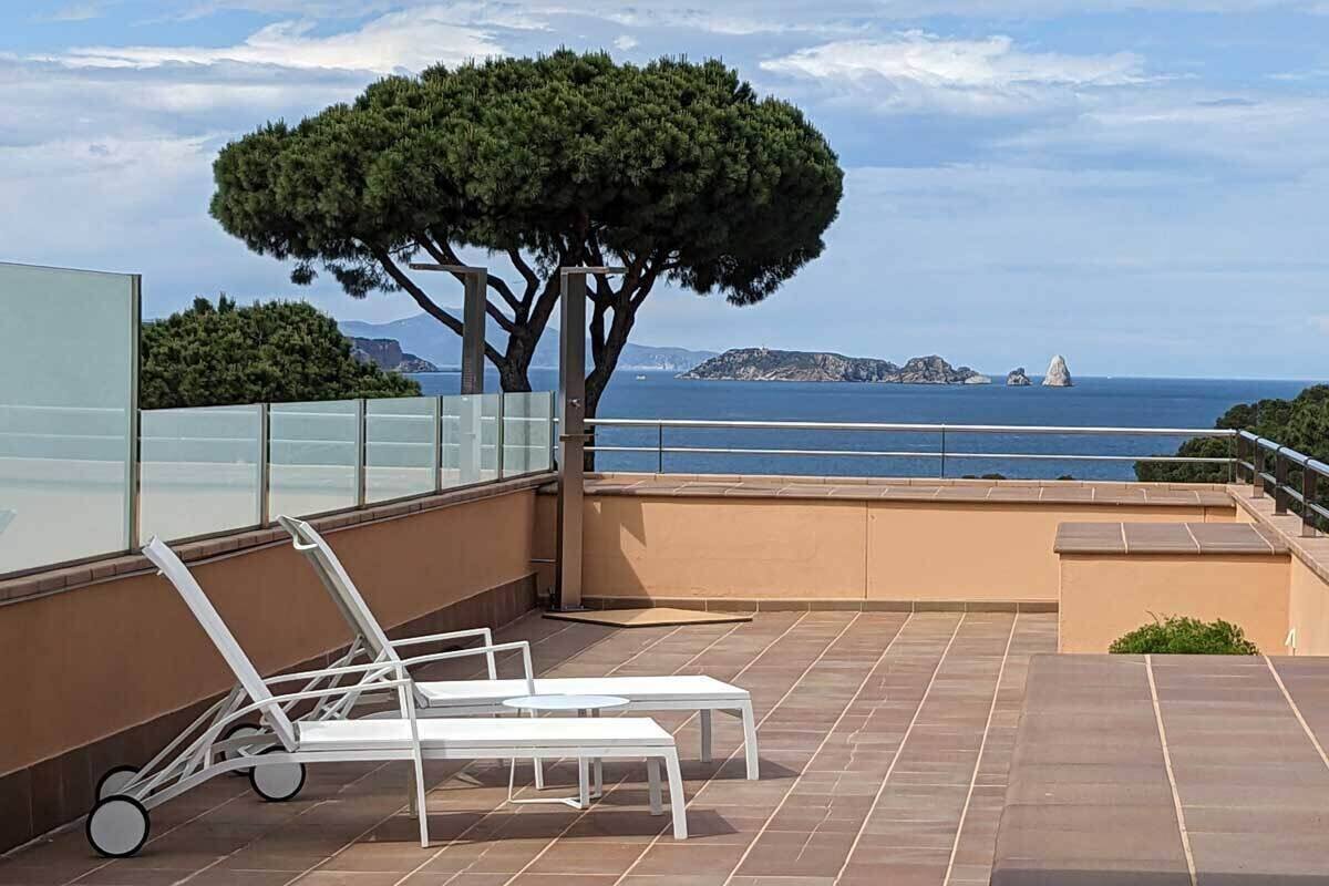 A rooftop terrace with two white lounge chairs, a glass balustrade, and a view of the sea and distant islands.