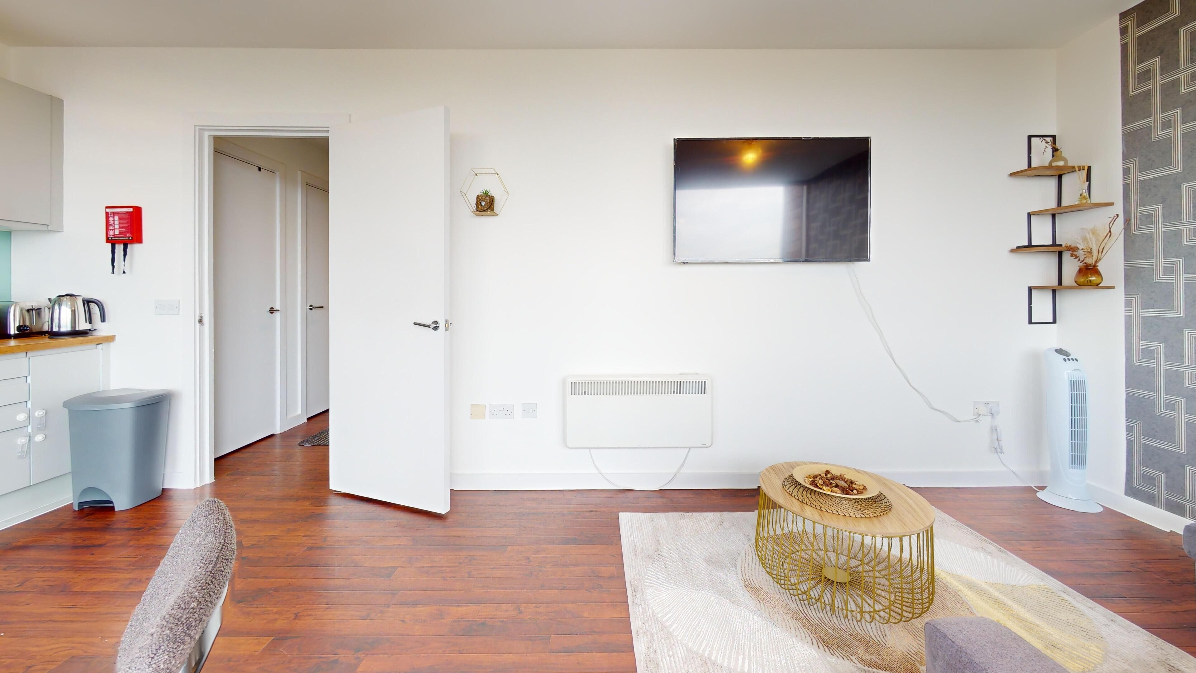 Un salon moderne avec un parquet, un radiateur blanc, un téléviseur fixé au mur et une petite table en bois sur laquelle repose un dessous de verre en fibres tressées.