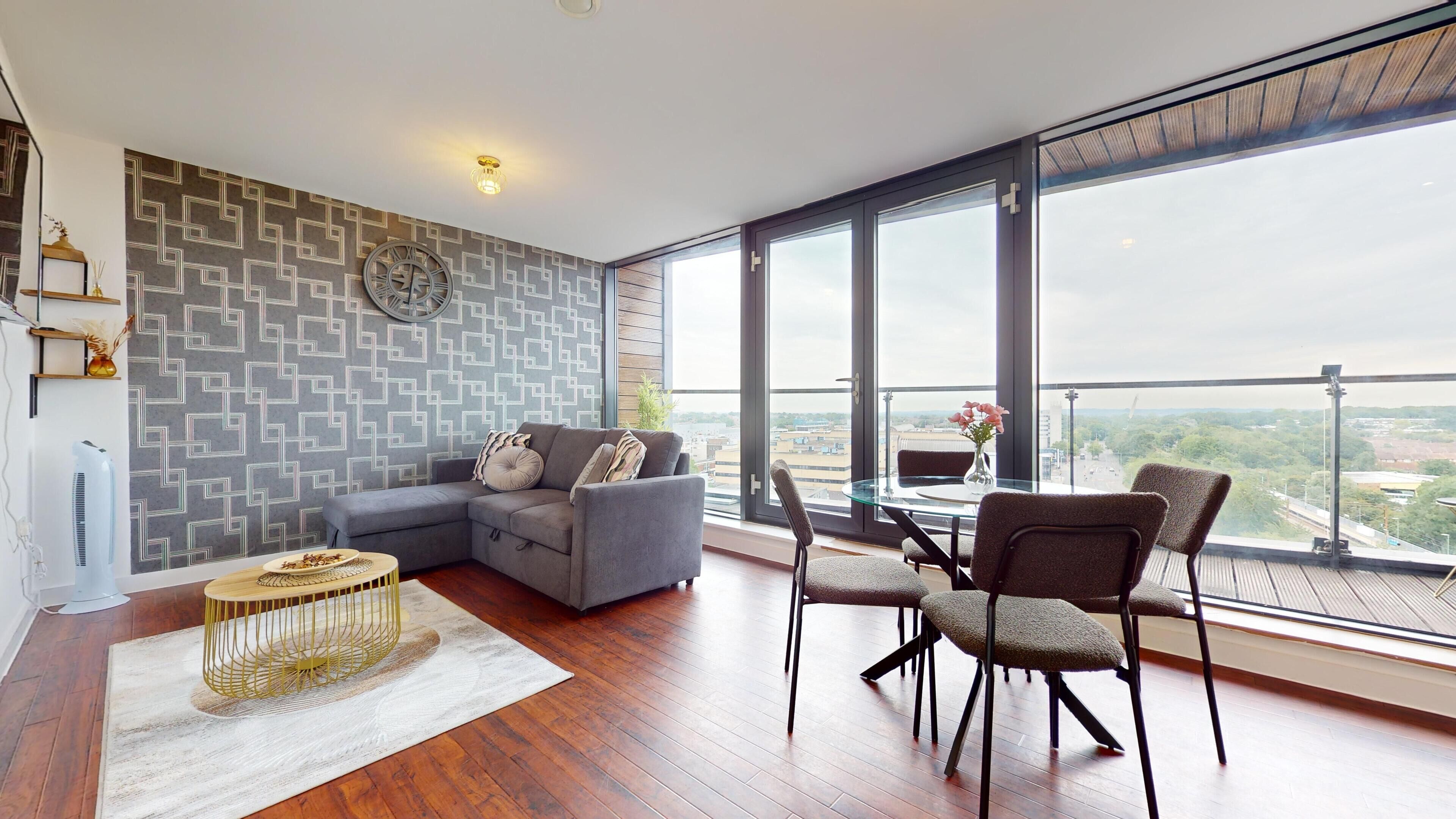 Una sala moderna con un sofá seccional gris, una mesa de comedor de vidrio con cuatro sillas, un ventanal con vista a la ciudad y una pared con diseño.