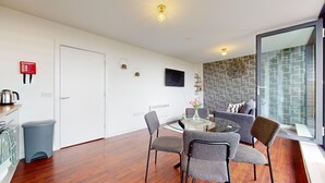 A modern living room with a glass dining table, grey upholstered chairs, a grey sofa, and a large window with patterned curtains. - Basildon - 2 Bed Flat - Sleeps 5 - Near Station (Basildon)