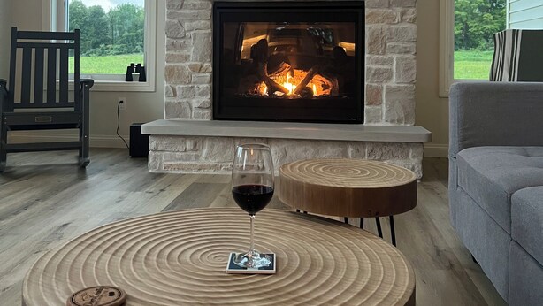 Una sala de estar con chimenea de piedra, un televisor de pantalla plana montado encima y una mesa de centro redonda de madera con una copa de vino tinto.