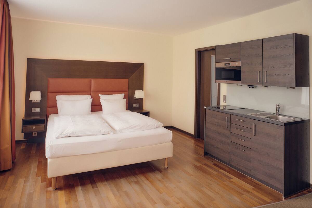 A hotel room with a bed, bedside tables, a built-in kitchen with cabinets and a microwave.