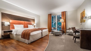 A hotel room with a large bed, two bedside tables, a desk, two chairs, a window with curtains, and a painting on the wall.
