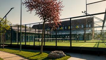 Instalación deportiva con cancha de tenis, un árbol y un edificio de fachada de vidrio.