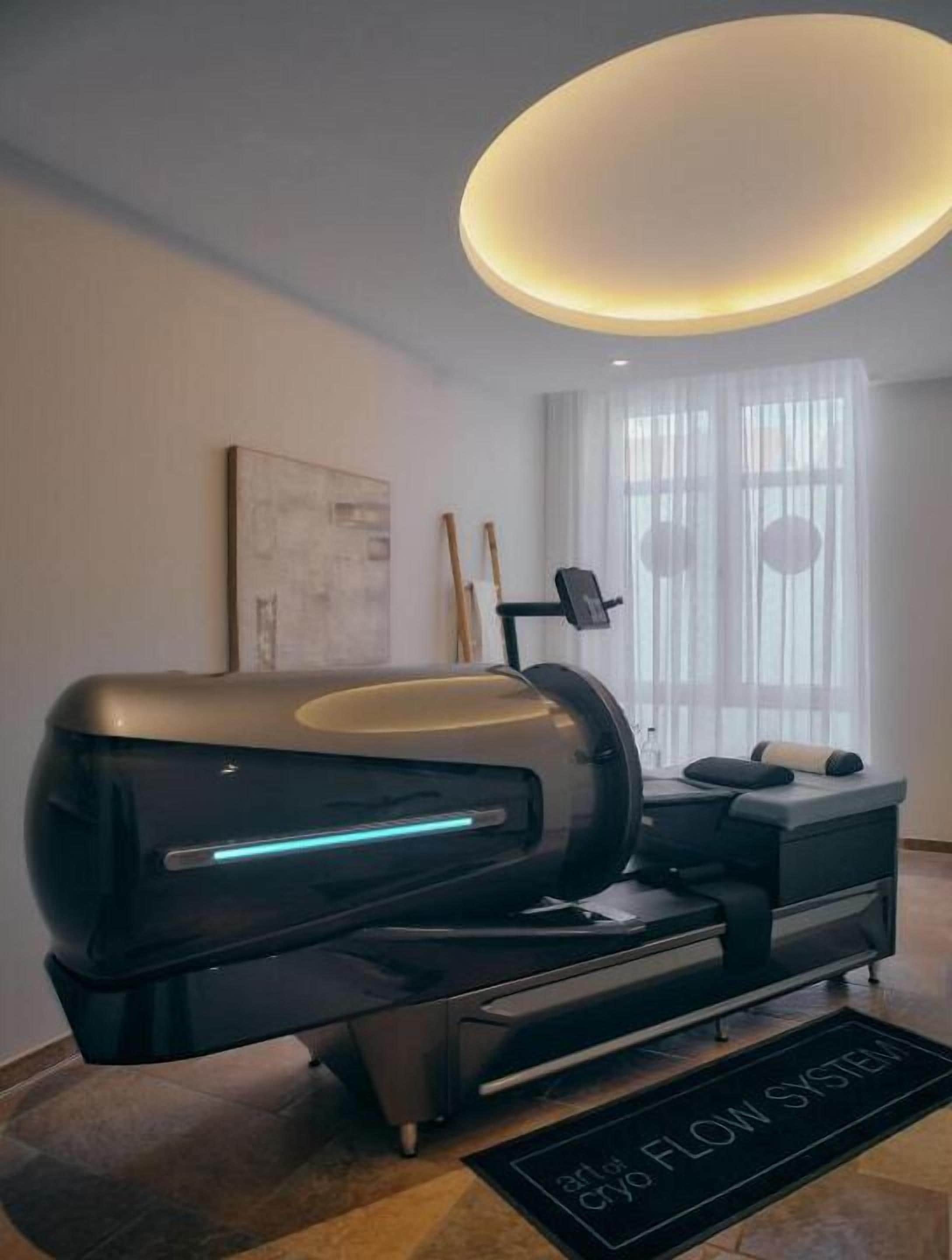 En modern massagefåtölj med en kontrollpanel, ett litet bord med en kudde och ett fönster med tunna, genomskinliga gardiner.