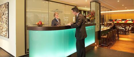 A reception desk with a glass partition, a fruit bowl, and a bar area with seating.