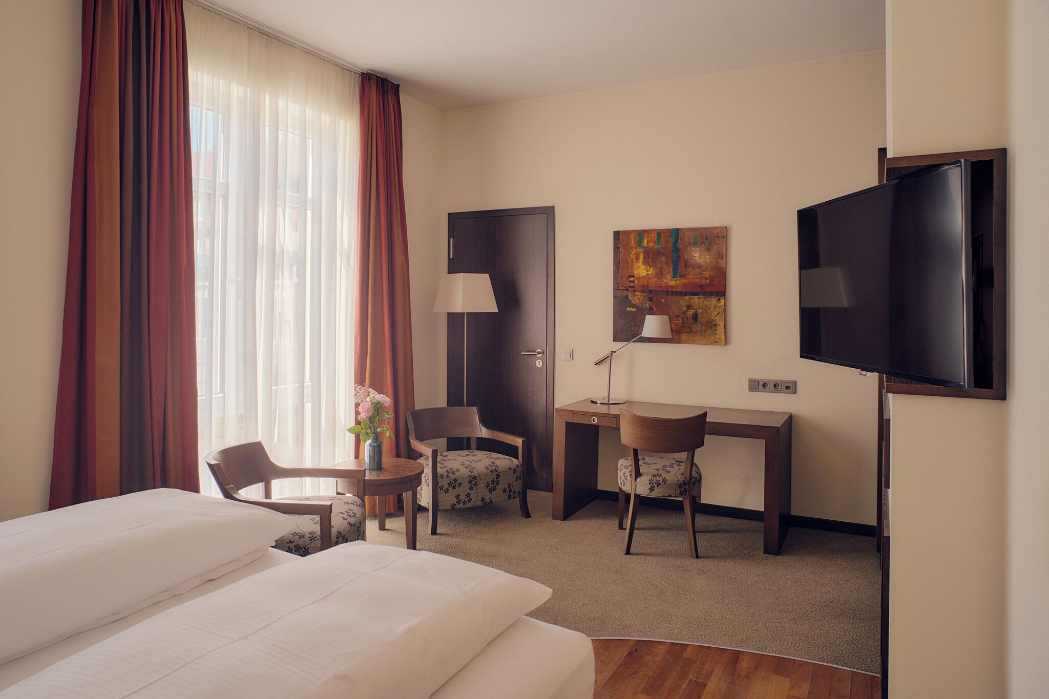 A hotel room with a bed, two chairs, a desk, a TV, and a painting on the wall.