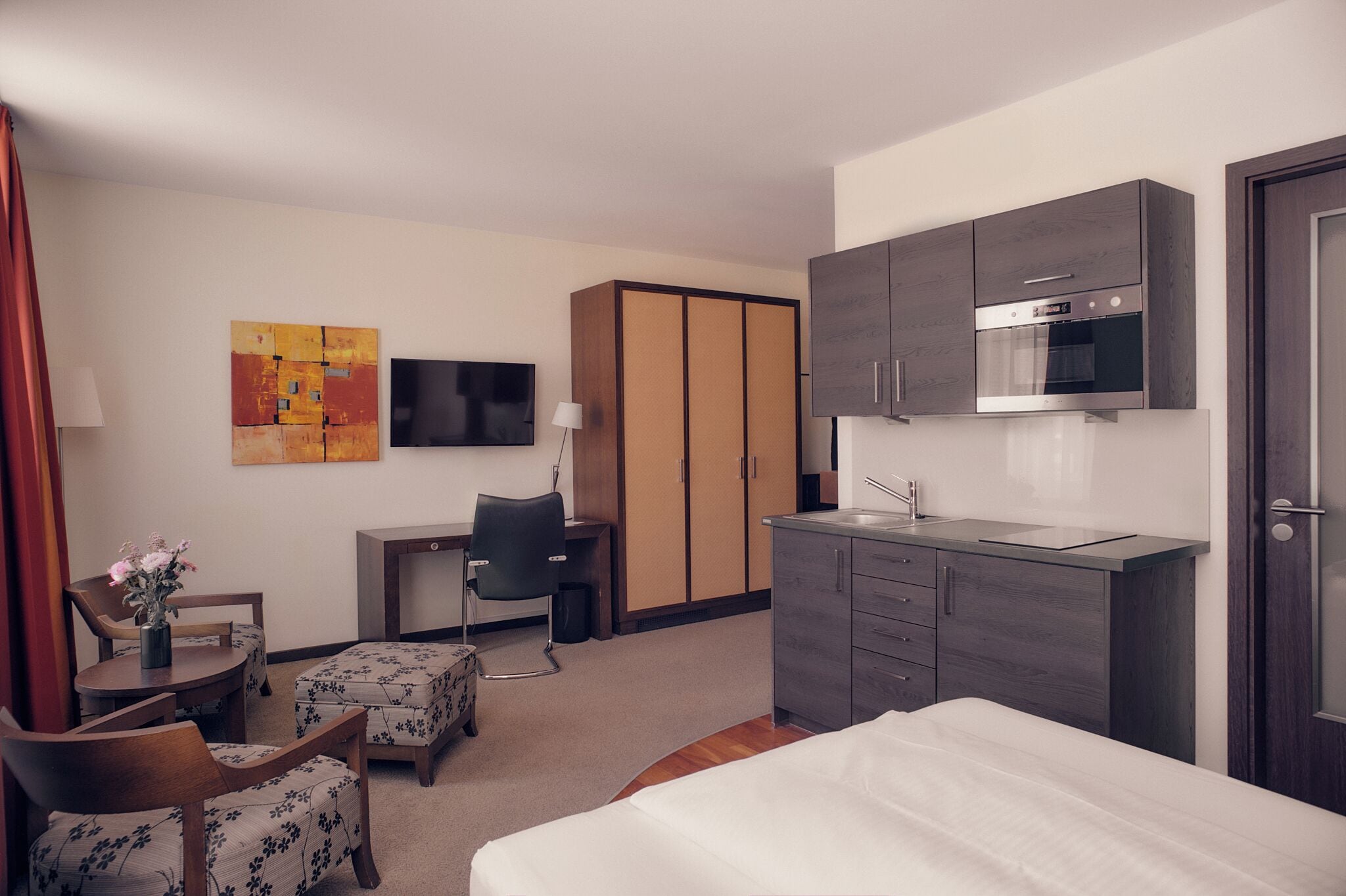 A hotel room with a bed, a desk with a chair, a TV, a kitchenette, and a wardrobe.