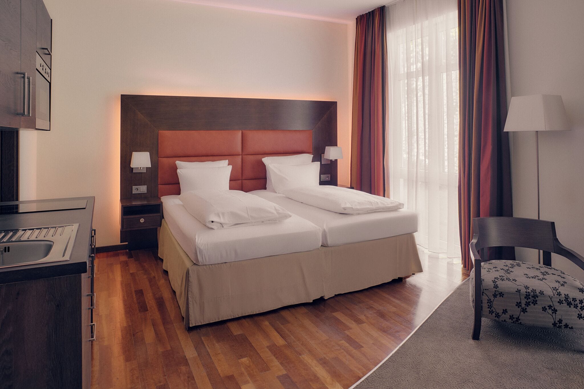 A hotel room with a double bed, bedside tables with lamps, a desk with a sink, a chair, and a large window with curtains.