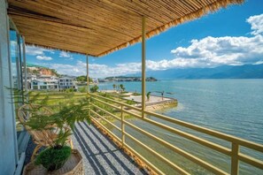 Un balcon offrant une vue dégagée sur un lac, un toit de chaume et une rive lointaine parsemée de bâtiments.