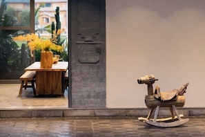 A wooden rocking chair and a table with a vase of yellow flowers in a room with a concrete pillar and a view of greenery outside.