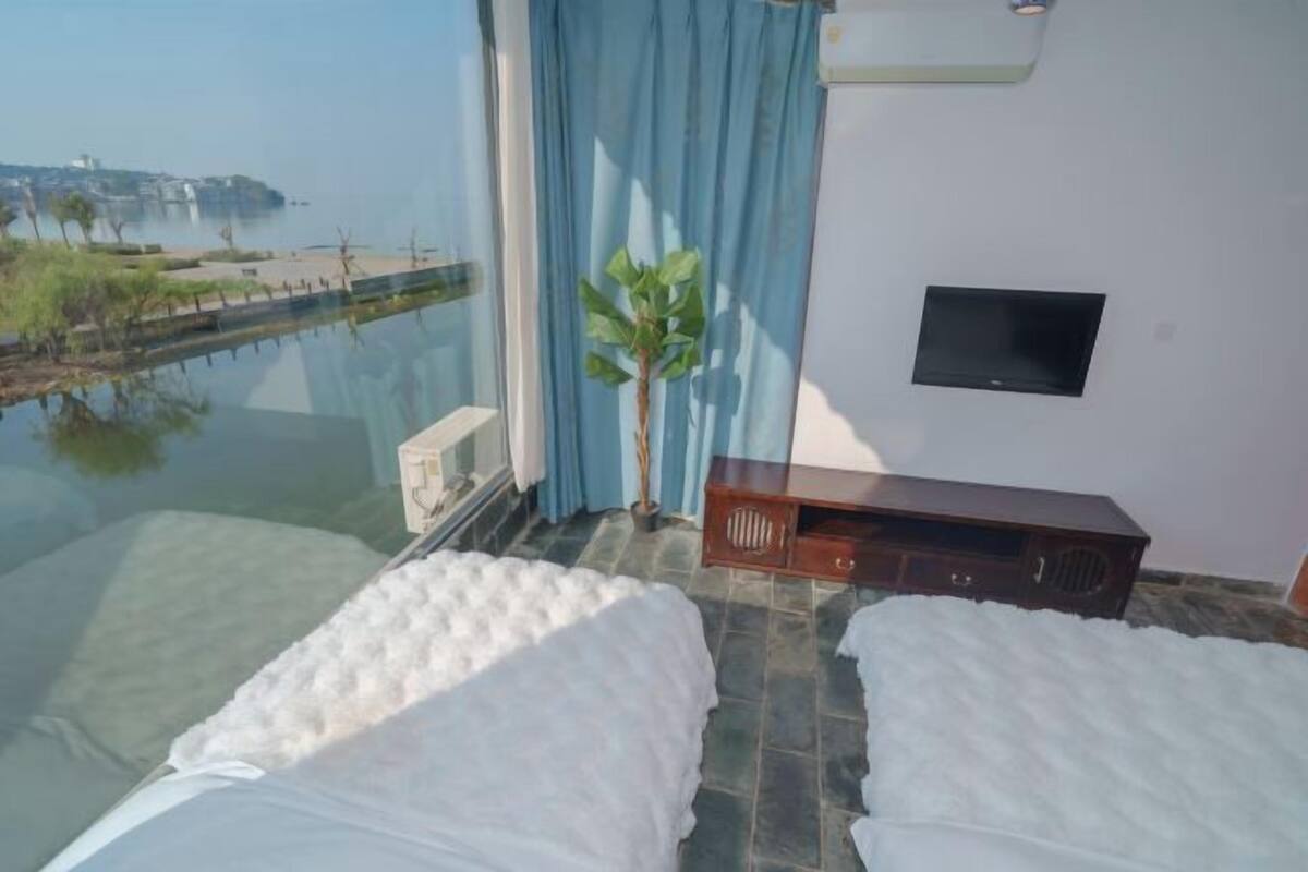 Una habitación con piso de vidrio, una cama, una planta pequeña, un televisor y vista a un cuerpo de agua.